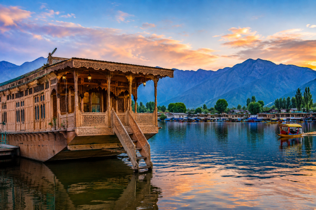 Traditional Kashmiri houseboat on Dal Lake in Srinagar at sunset with shikaras and Himalayan mountains in the background.